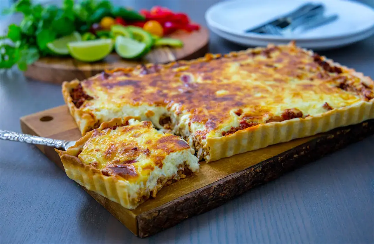 En gyllene tacopaj med köttfärs och ost, redo att serveras. Tillbehör som lime och koriander syns i bakgrunden.