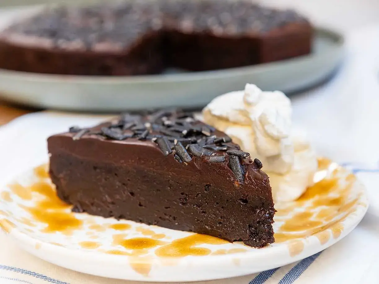 En saftig kladdkaka med lakritsströssel, serverad med vispad grädde och en skvätt sirap på en mönstrad tallrik.