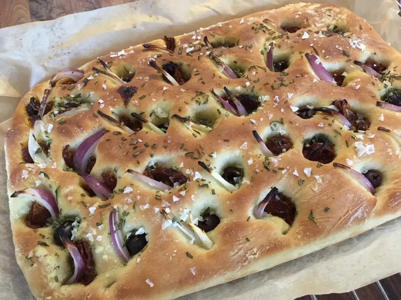 En gyllene focaccia l&aring;ngpanna med r&ouml;dl&ouml;k, oliver och soltorkade tomater, toppad med &ouml;rter och flingsalt.