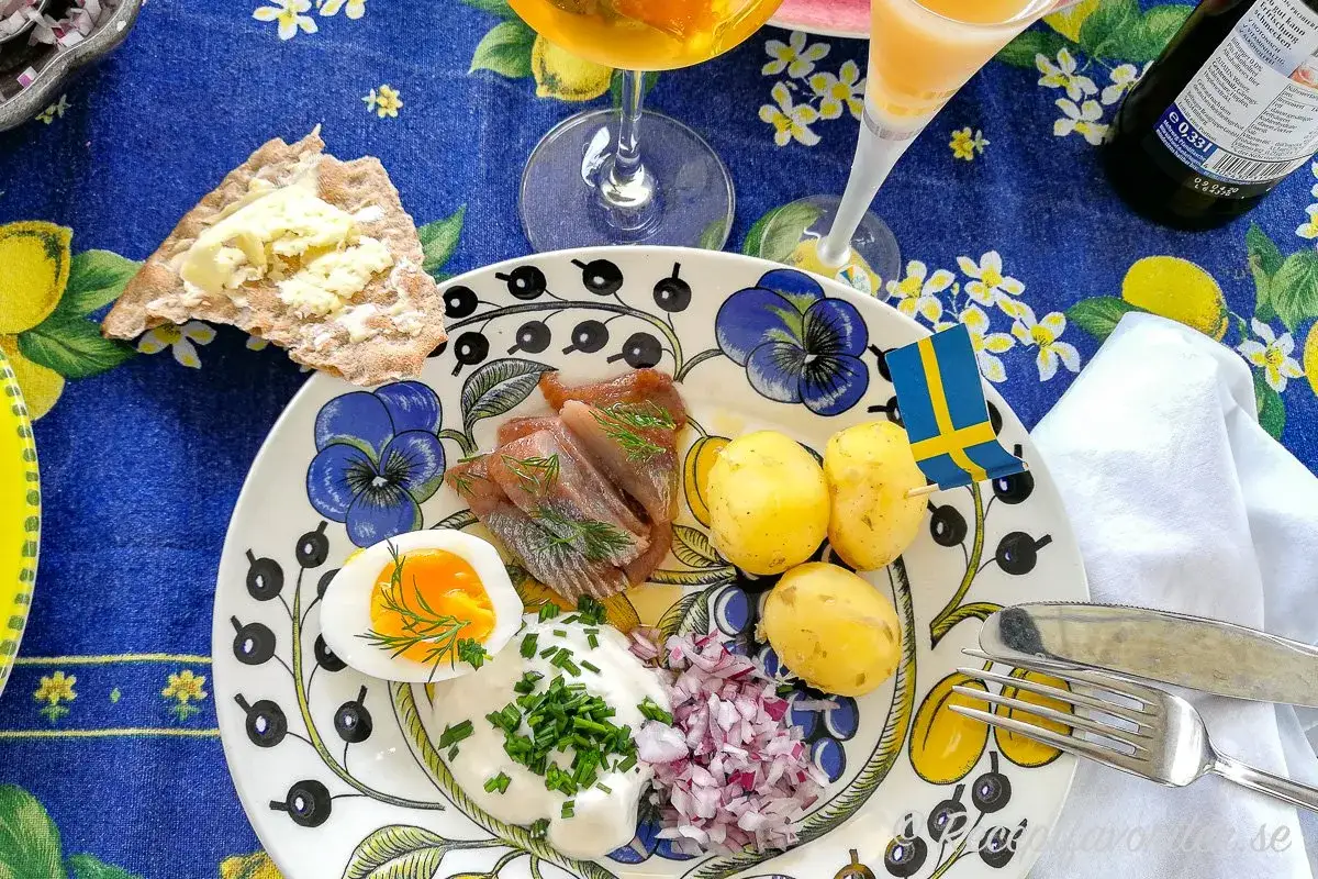 En tallrik med klassisk midsommar mat: sill, färskpotatis, ägg och gräddfil. En svensk flagga vajar stolt.