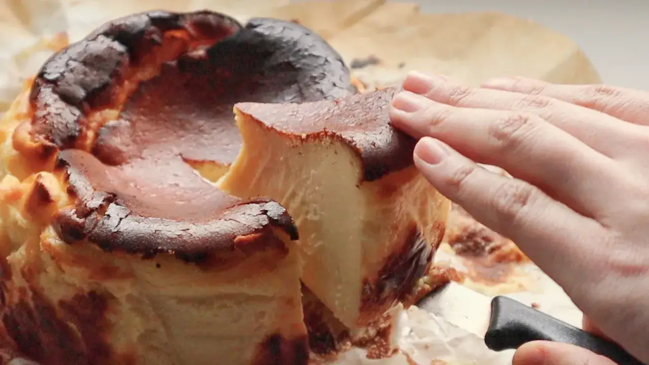 En hand skär en bit av en mörk, karamelliserad baskisk cheesecake med en krämig insida.