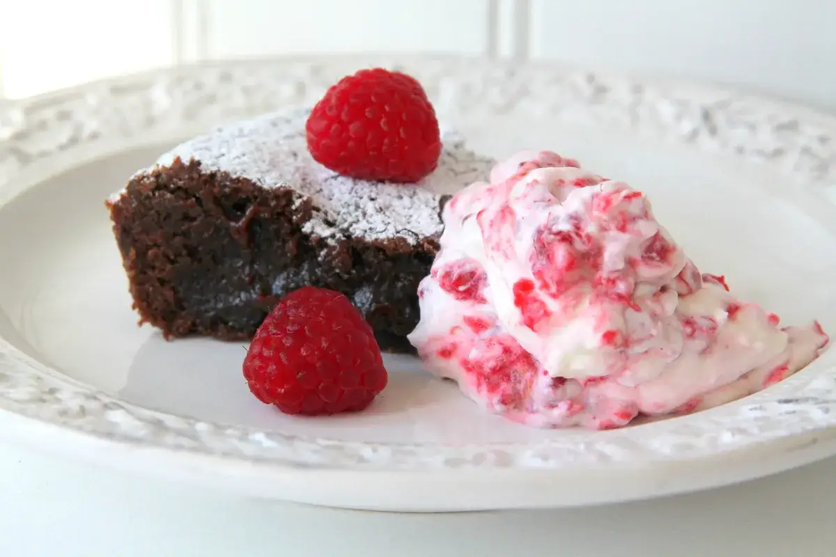 En bit kladdkaka med lakrits, pudrad med florsocker och garnerad med hallon. Serveras med en klick hallongr&auml;dde.