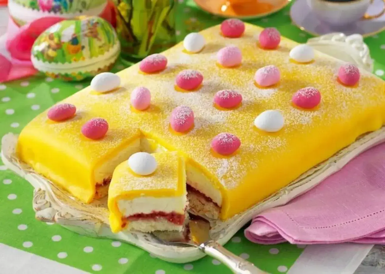 En gul påsktårta enkel, dekorerad med rosa och vita marsipanägg, pudrad med florsocker. En bit är uppskuren och visar lager av kräm och sylt.