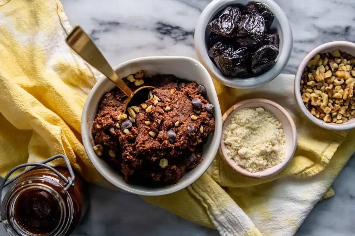 Chokladig cookie dough recept med nötter och chokladknappar. Ingredienser som katrinplommon, valnötter och mandelmjöl syns bredvid.