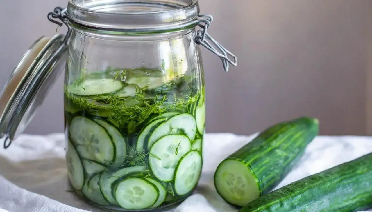 En burk med skivade gurkor och dill, redo för ett saltgurka recept. Två hela gurkor ligger bredvid.