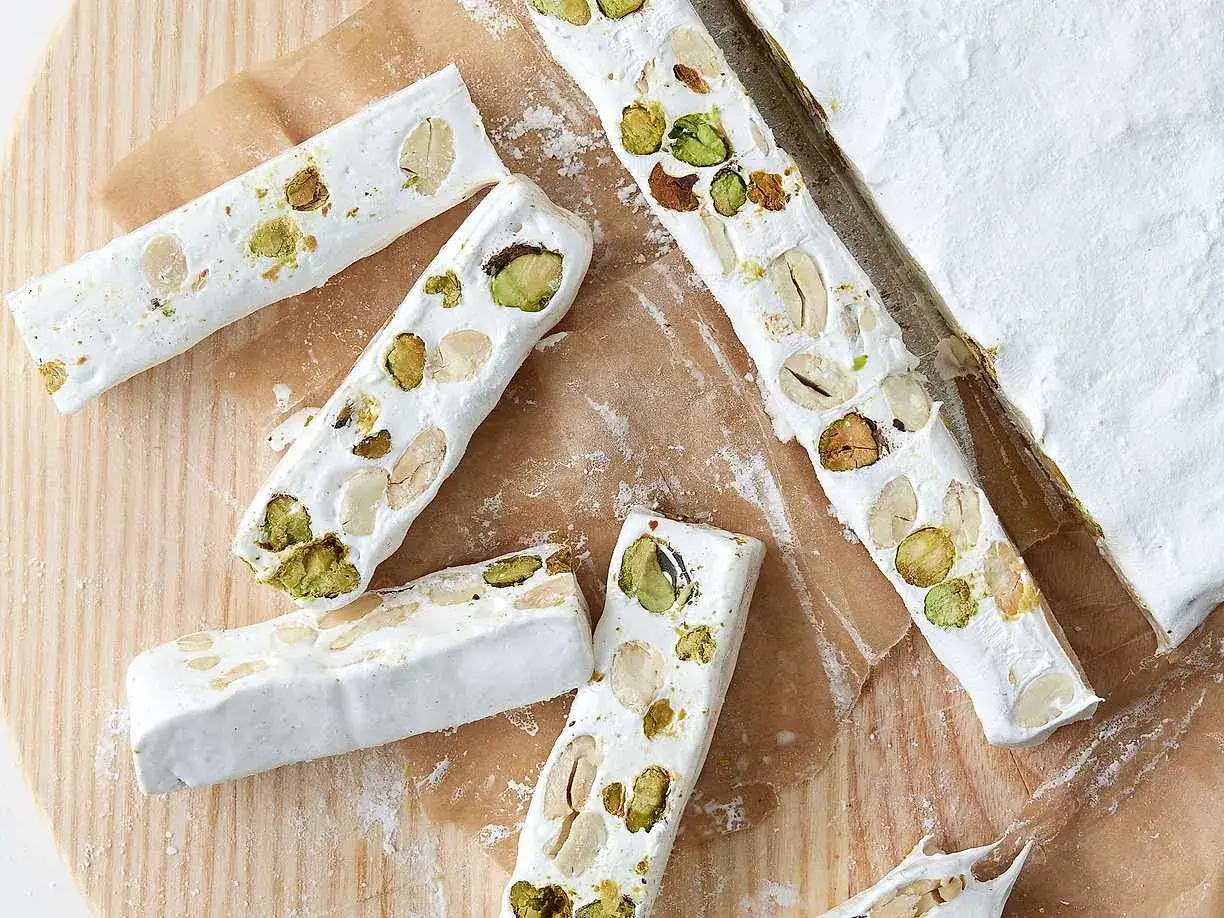 Flera bitar fransk nougat med pistagenötter och mandlar ligger utspridda på ett träunderlag.