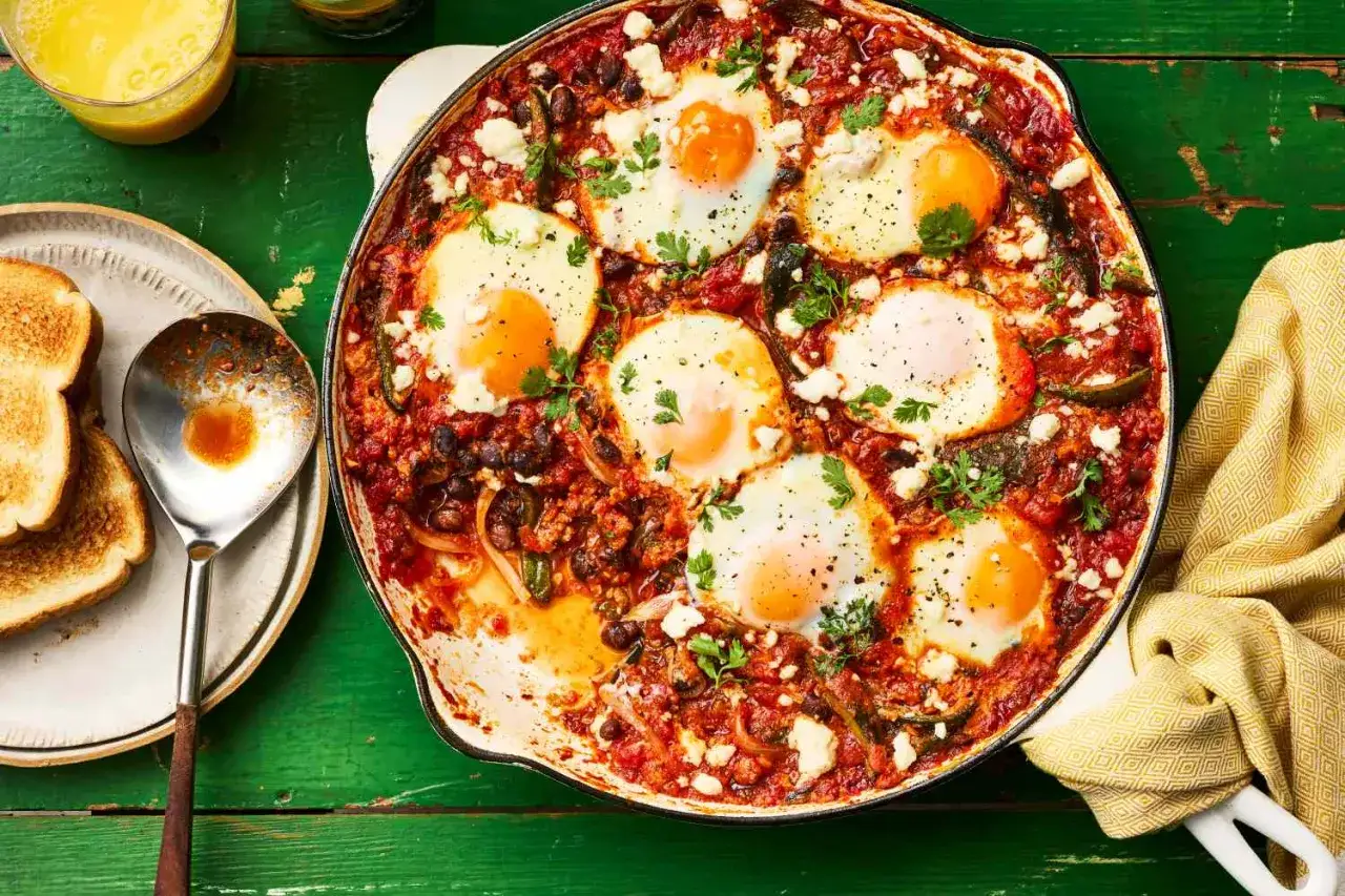 En gjutjärnspanna fylld med en smakrik huevos rancheros-röra med bönor, tomater och sex stekta ägg, toppad med ost och persilja. Bredvid syns rostat bröd.